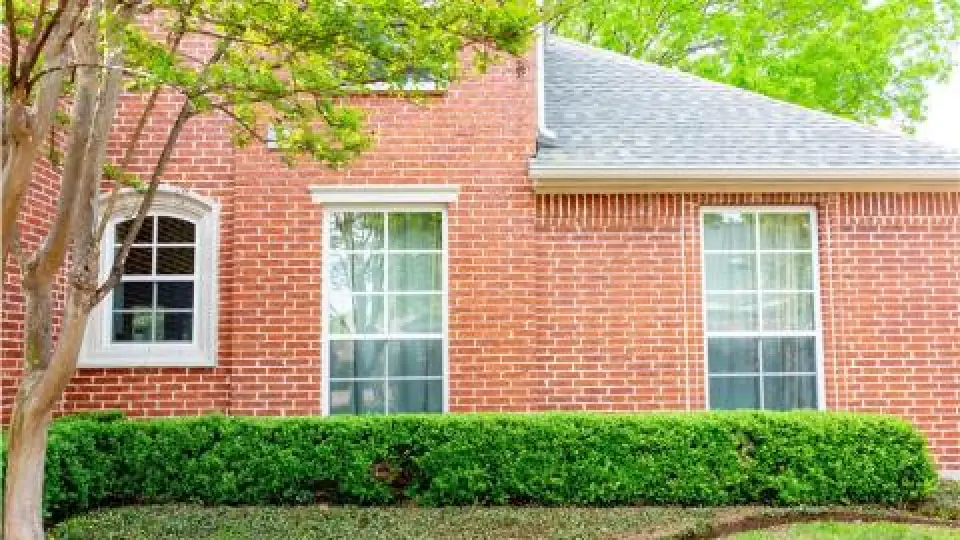 Red brick house with three white-trimmed windows and green bushes in front.