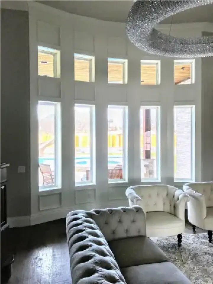 Tall white windows and tufted chairs in a bright, elegant living room with a modern chandelier.
