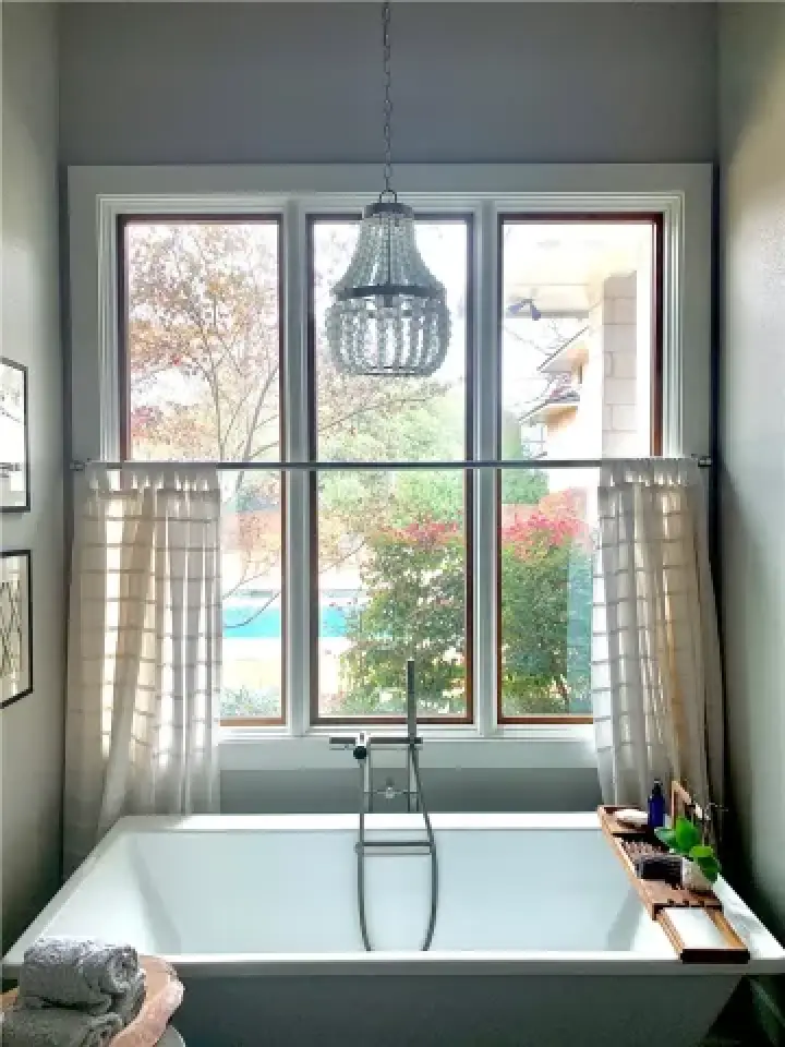 Modern bathroom with a large window, soaking tub, white curtains, and a beaded chandelier.