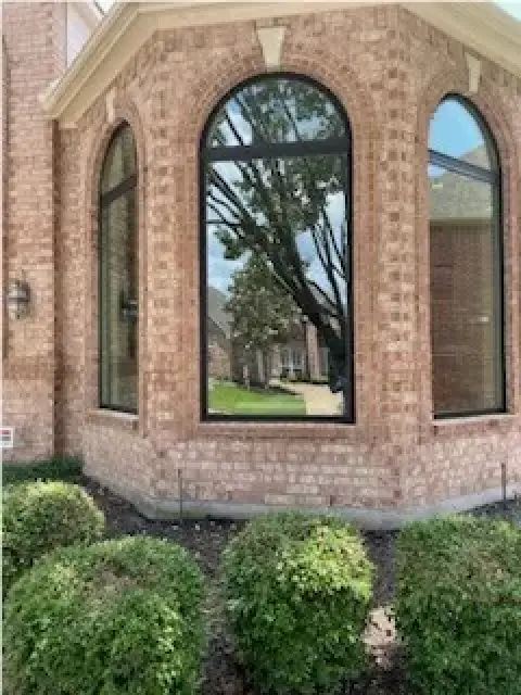 Large arched windows on a brick house reflect trees and nearby homes, with bushes in front.