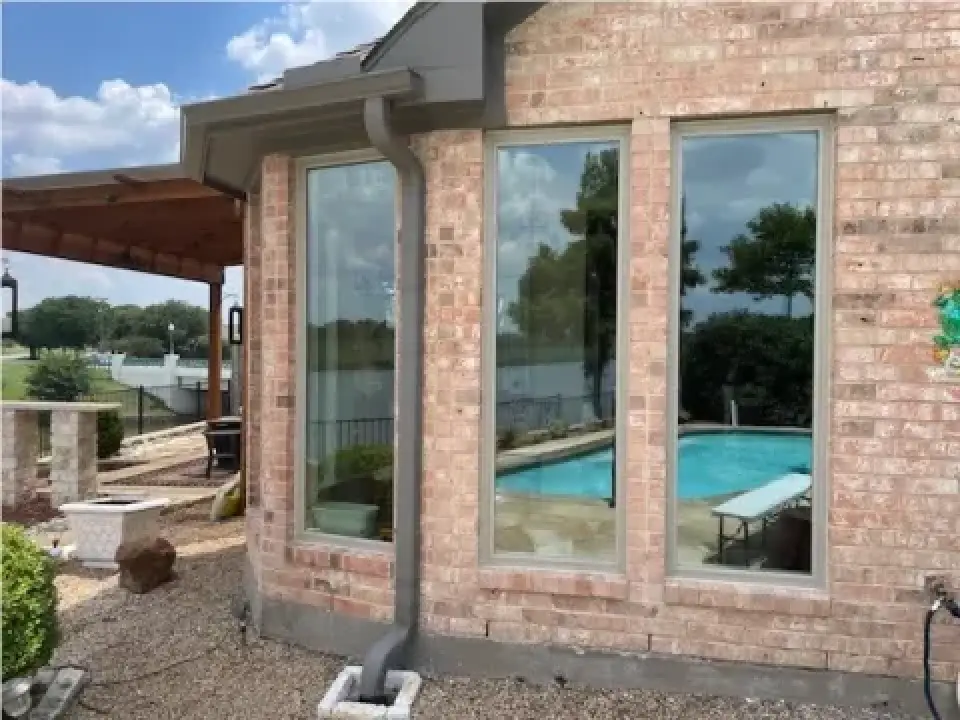 Brick house with large windows reflecting a swimming pool, trees, and a partly cloudy sky.