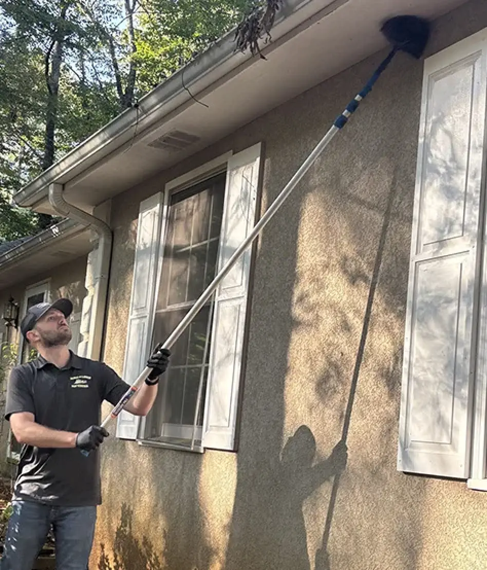 Pest control technician removing spider webs from a home's eaves.