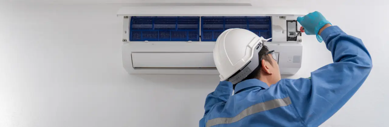 Technician in blue uniform and helmet repairing a wall-mounted air conditioning unit.