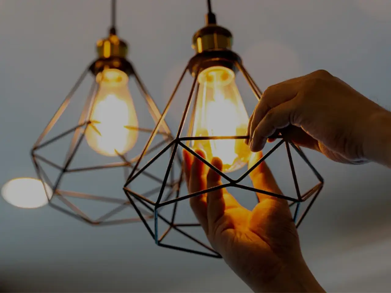 Hands adjust a geometric pendant shade around a glowing light bulb.