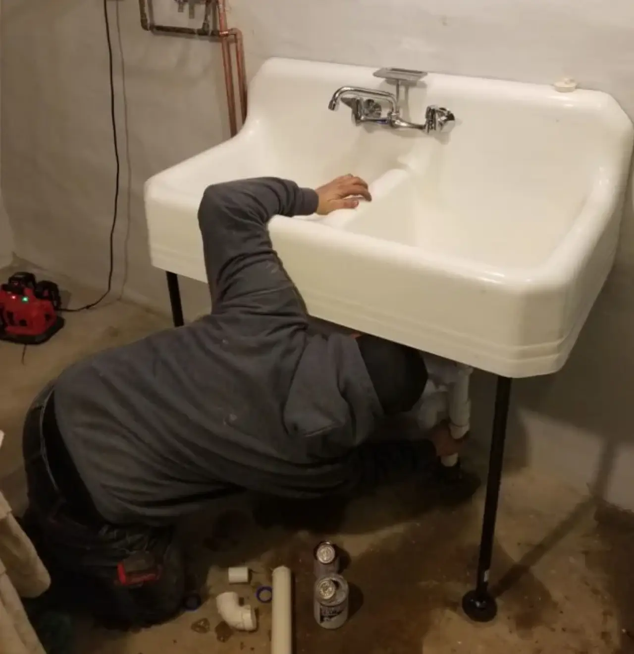 A person repairs pipes under a white sink; tools and cans are on the floor nearby.