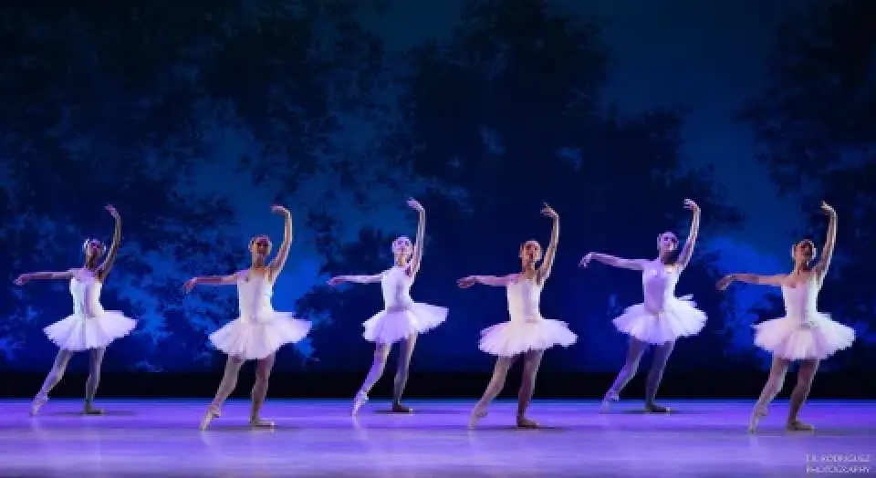 Six ballerinas in white tutus perform on a blue-lit stage.