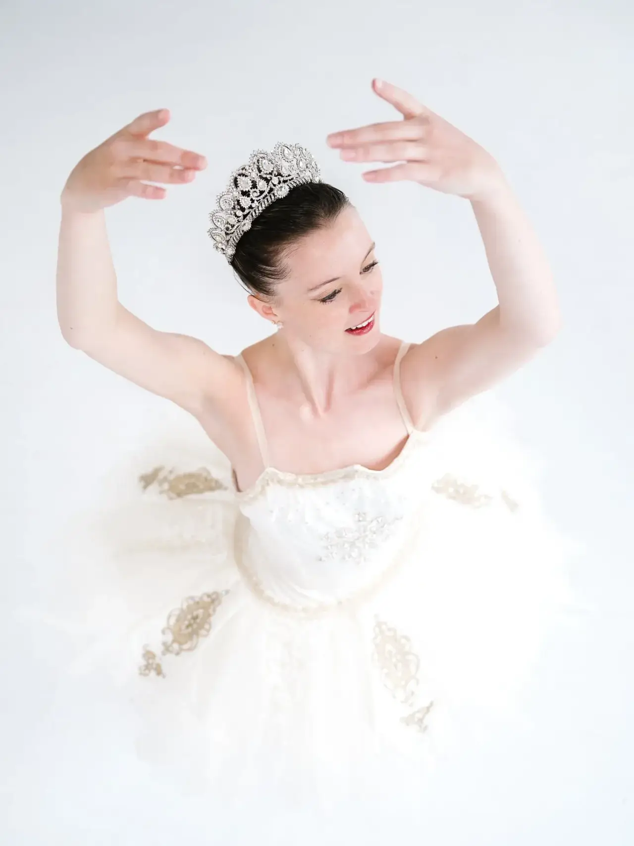 Ballerina in a white costume and tiara with raised arms.