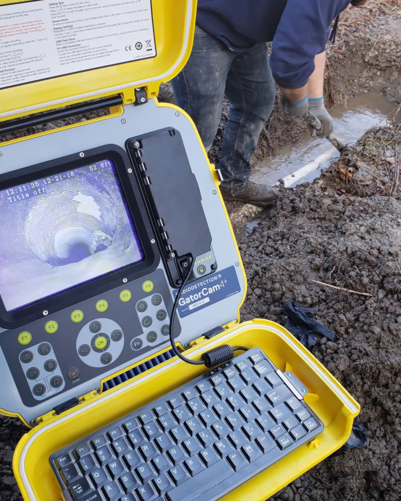 A worker checks a pipe near mud while a camera displays the inside view of the pipe.