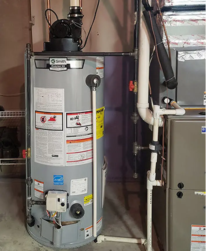 A gray water heater stands next to a furnace in a basement with visible pipes and labels.