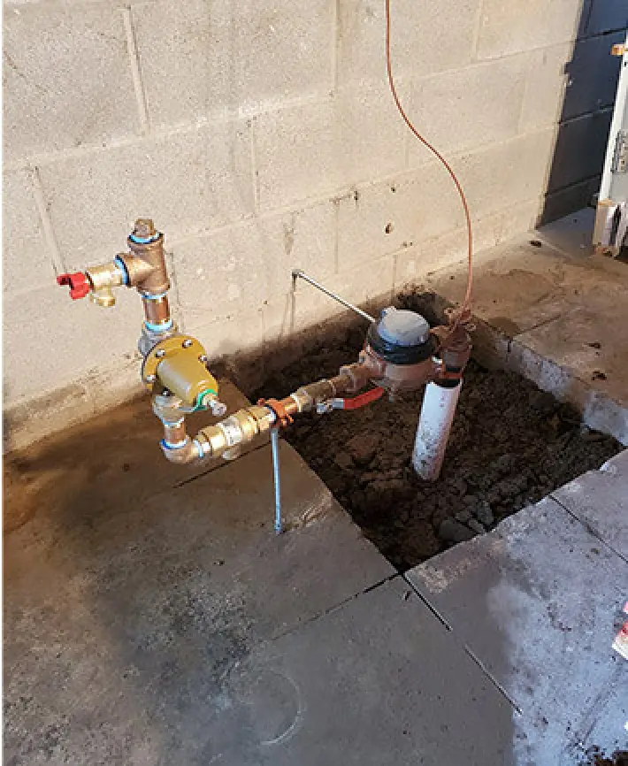 A water meter with connected pipes, installed by Palmyra plumbers, sits by a basement floor hole.