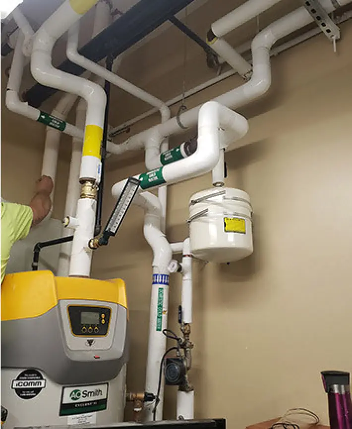 White pipes and a water heater system mounted on a beige wall, with a person's arm visible.