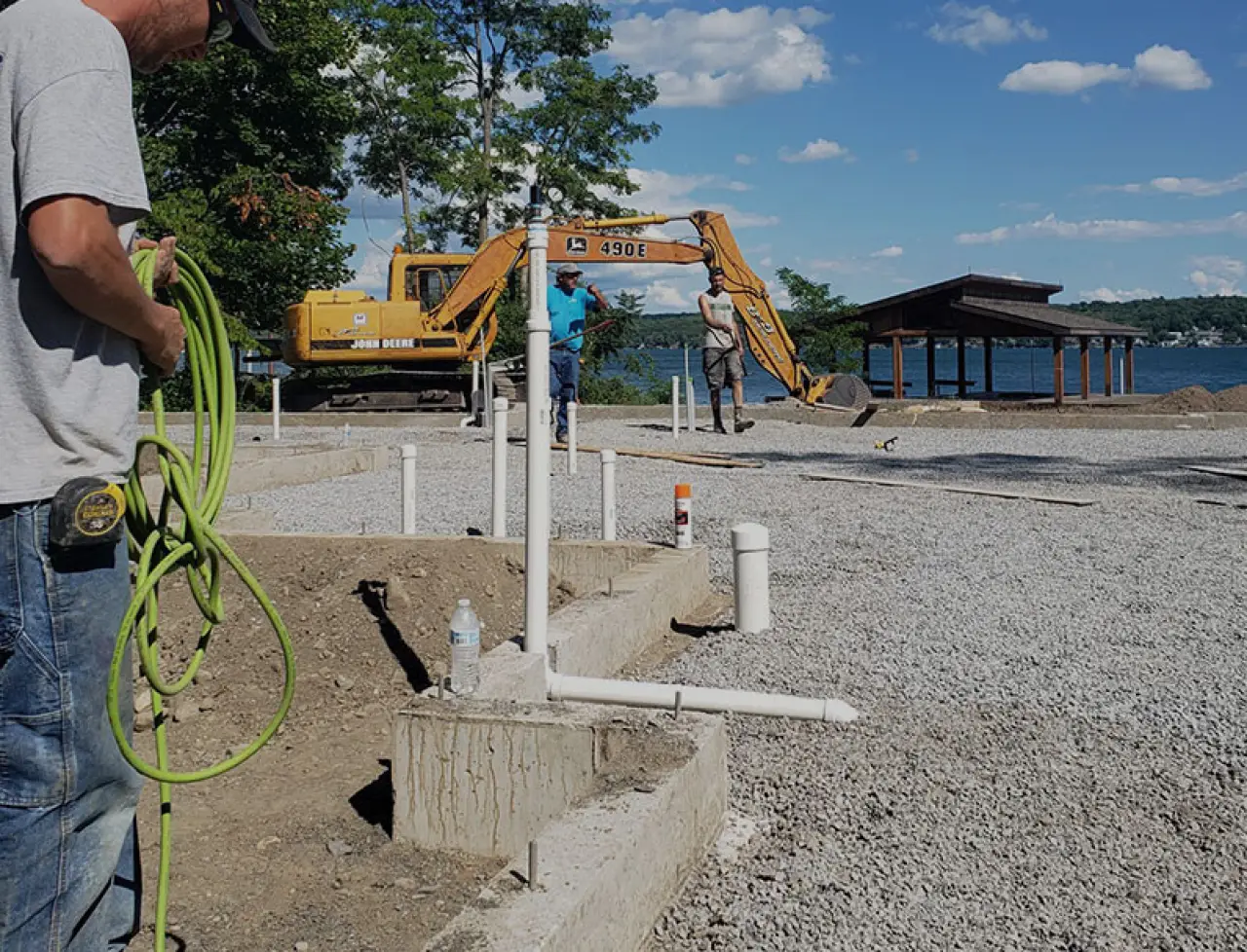 Three workers at a lakeside construction site with pipes, gravel, and an excavator.