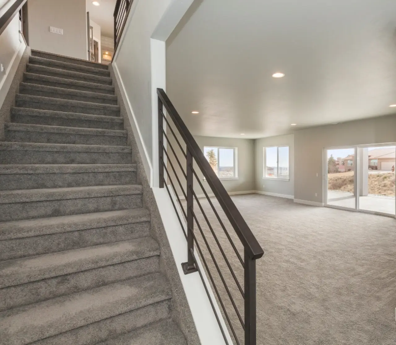 Carpeted stairs beside a large, empty room with windows and neutral-colored walls and flooring.