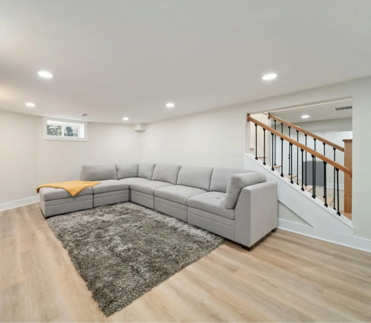 Basement room with gray sectional sofa, shaggy rug, wood stairs, and recessed lighting.
