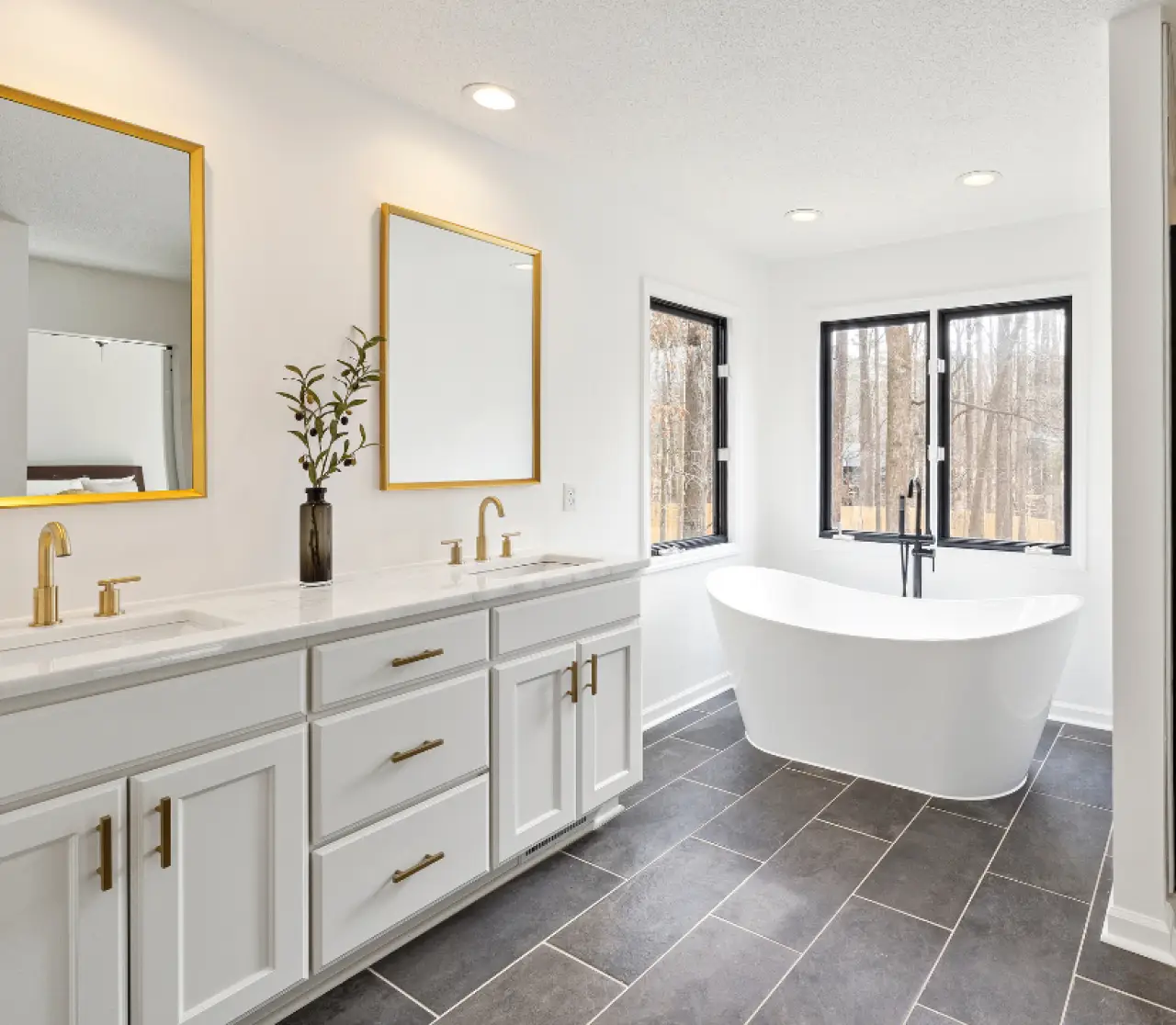 Modern white bathroom with a freestanding tub, double vanity, gold fixtures, and large windows.