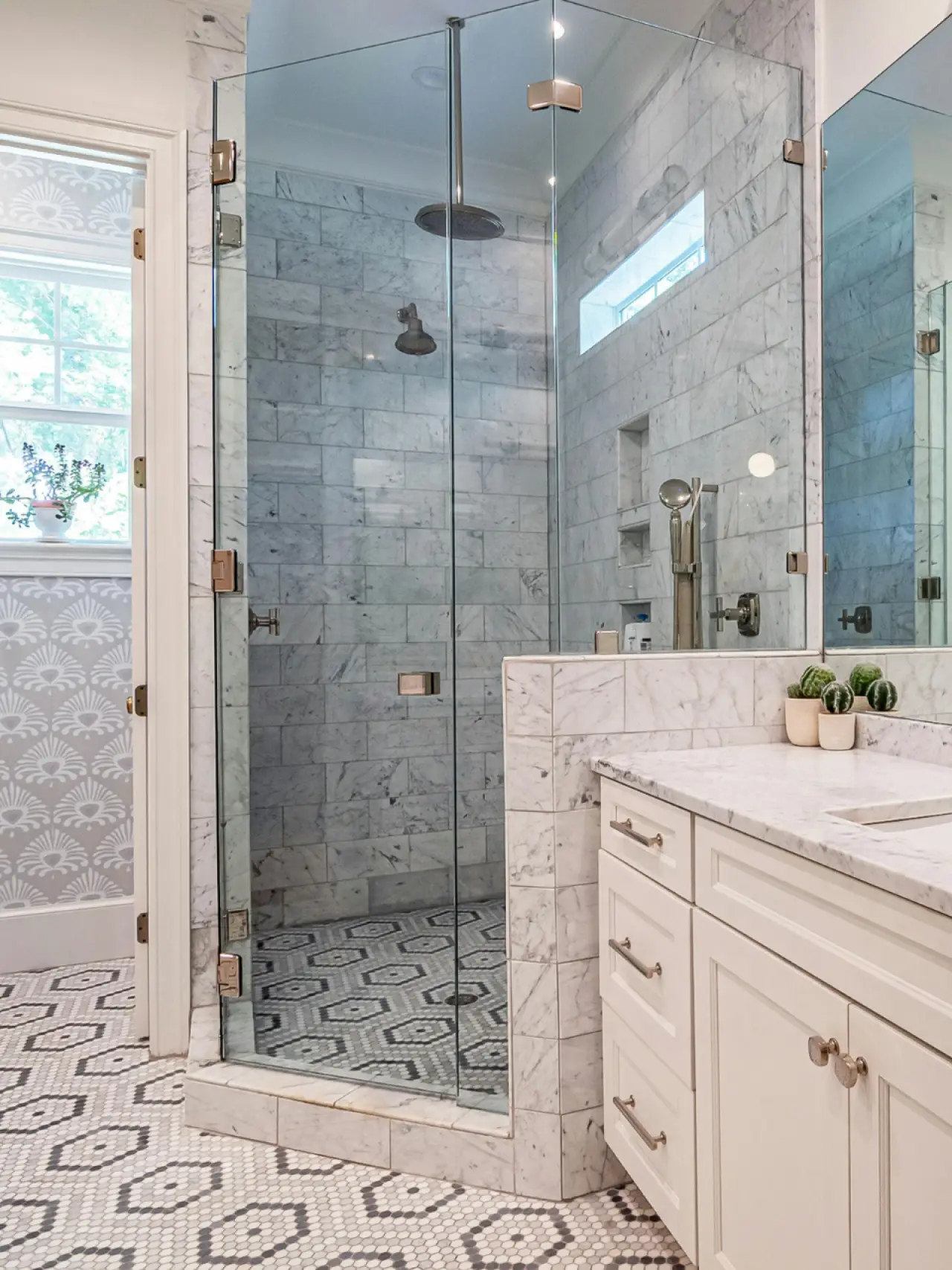 Marble bathroom with glass shower, white vanity, hexagon tile floor, and small potted plants.