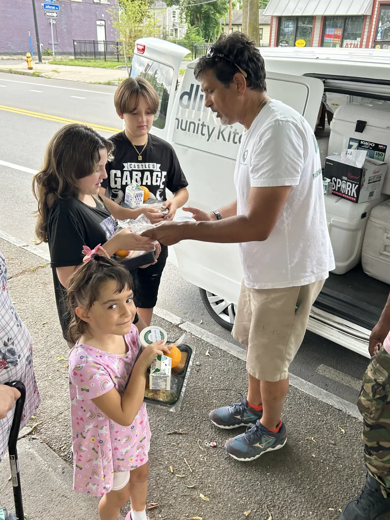 A man hands three children food for Summer Meals as a Foodlink truck is parked in the background