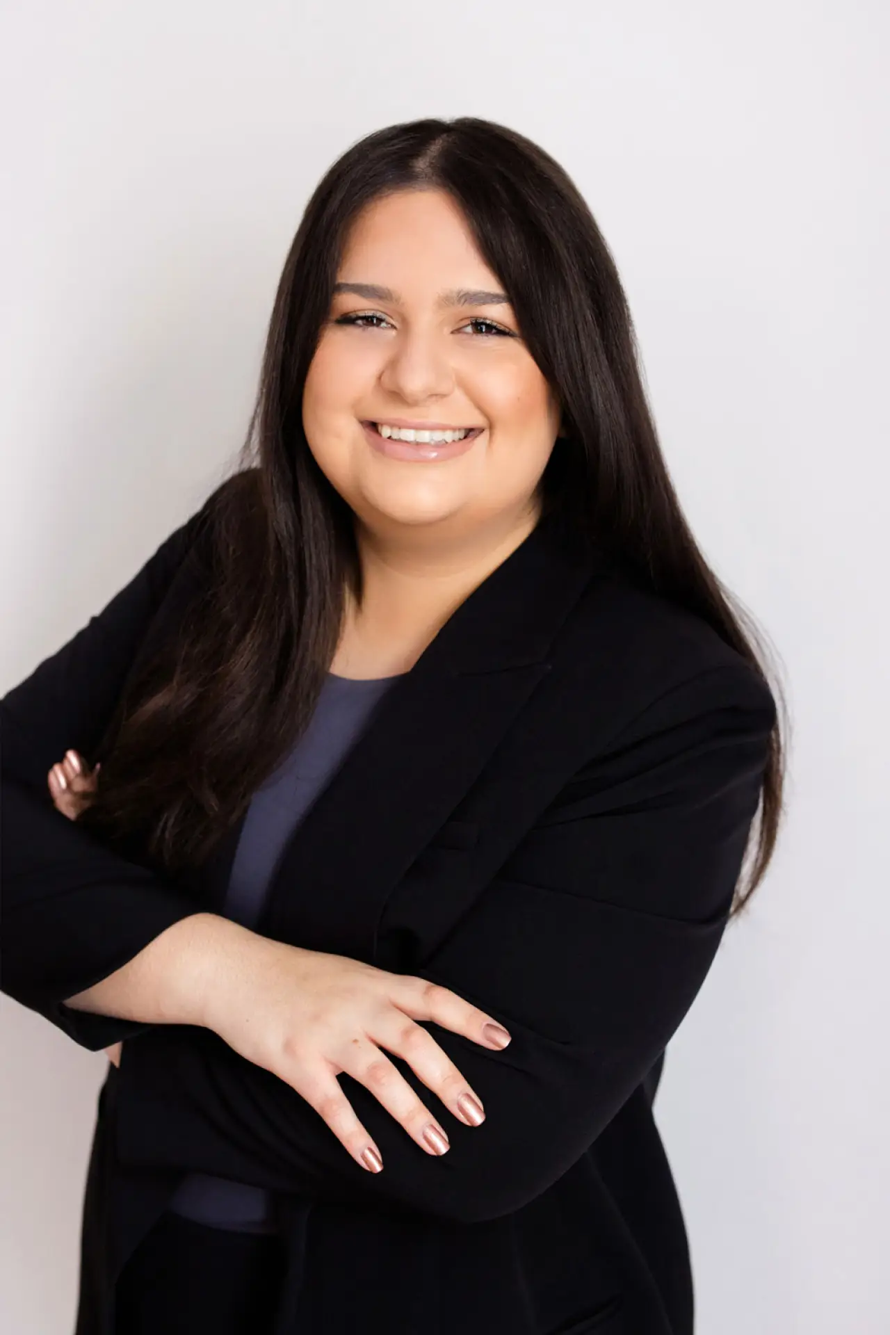 Smiling woman with long dark hair in a black blazer, arms crossed, on a light background.
