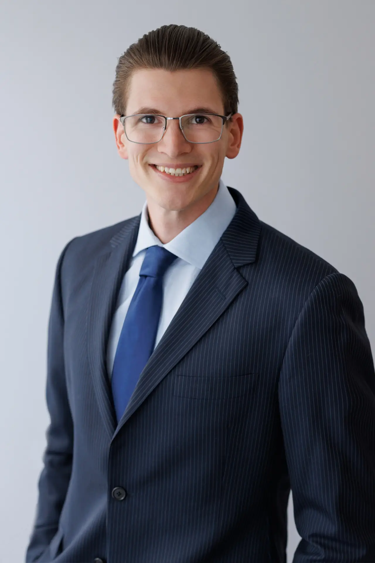 Smiling man in a dark striped suit, blue tie, and glasses against a light background.
