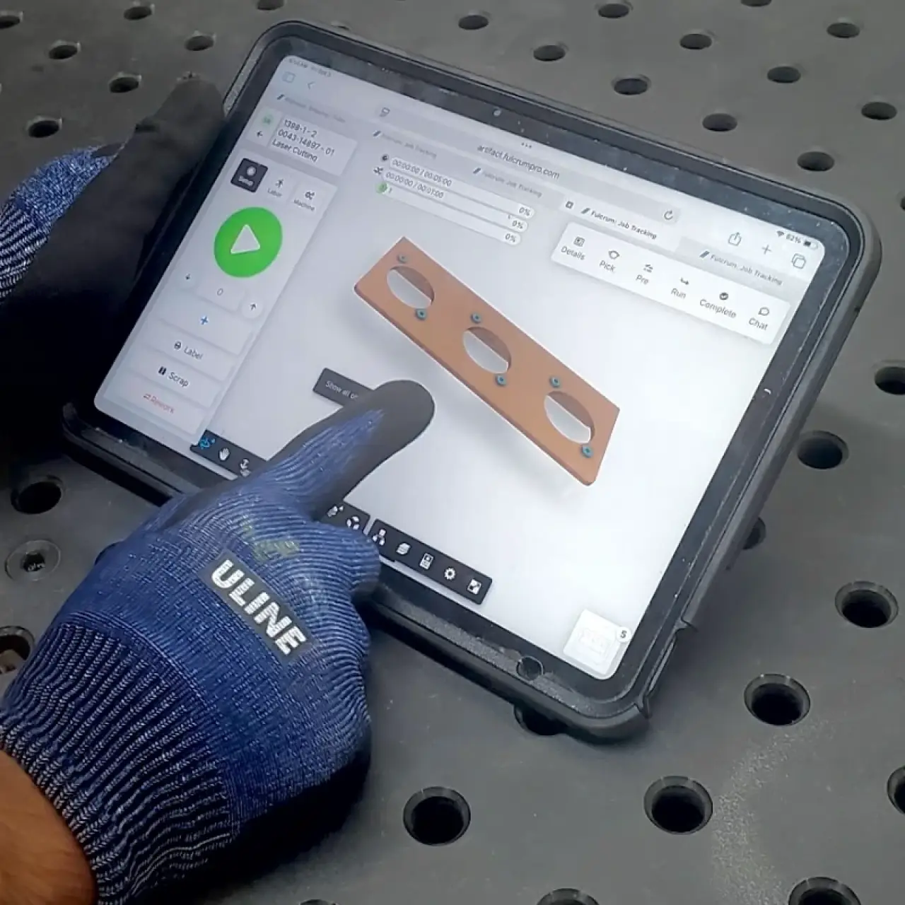 Gloved hands operate a tablet displaying a 3D model of a metal part on a perforated surface.
