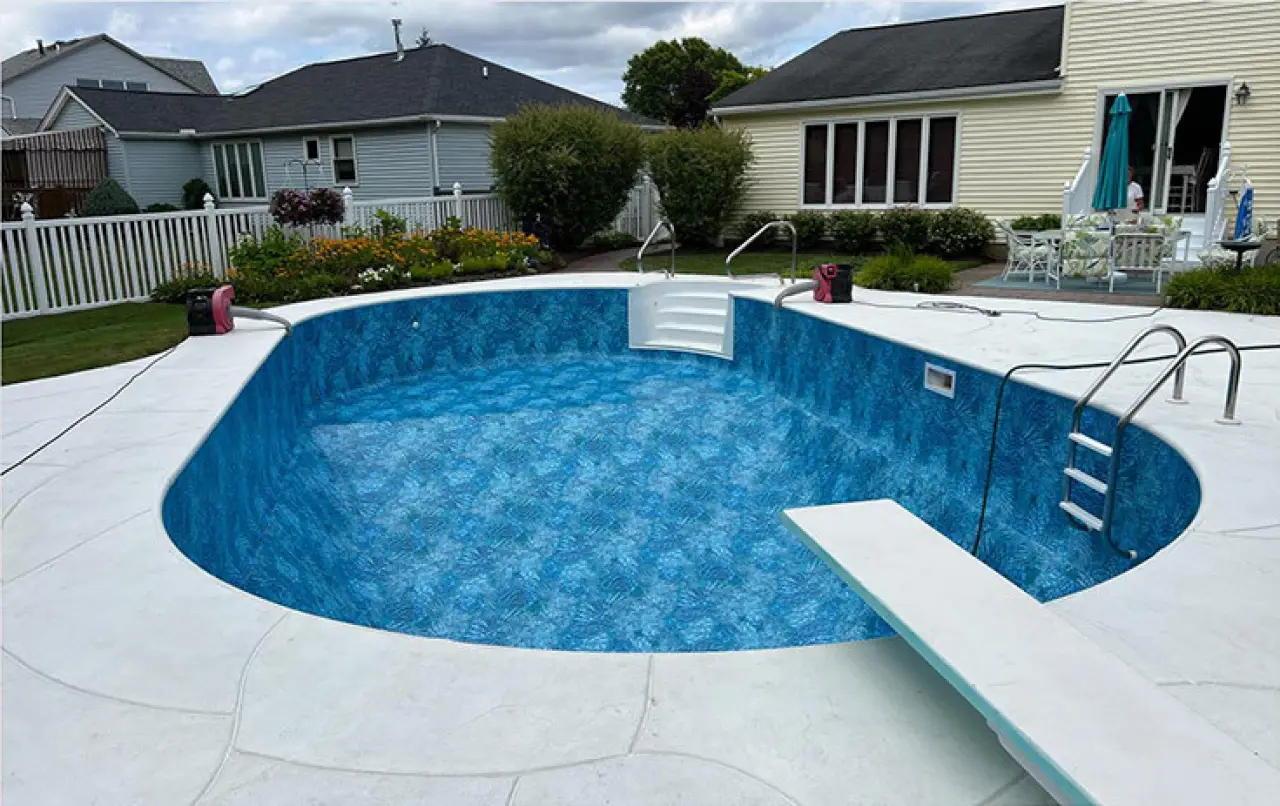 Empty in-ground swimming pool in a backyard, surrounded by patio, with a diving board and steps.