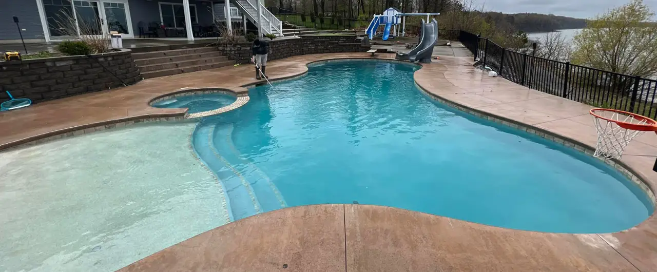 A backyard pool with a hot tub, slide, basketball hoop, and a person cleaning the area.