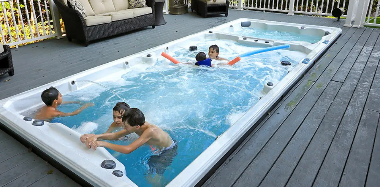 Four children play with pool noodles in a large outdoor hot tub on a wooden deck.