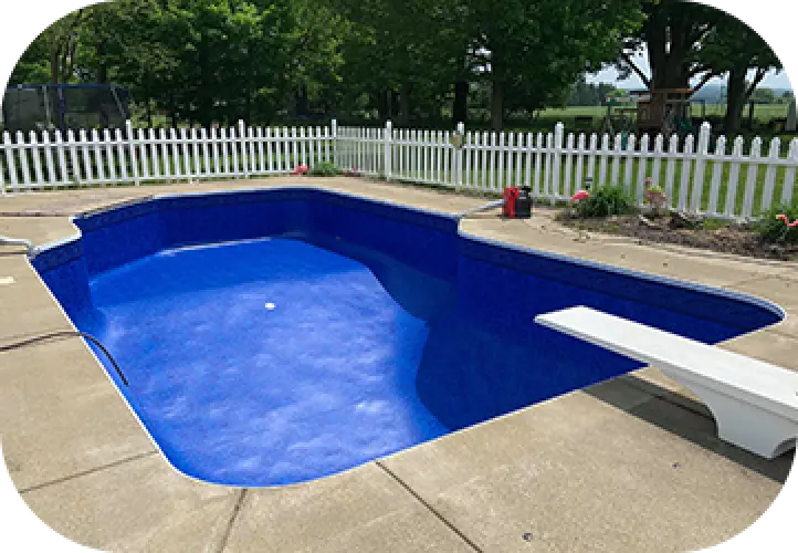 Small backyard swimming pool with a diving board, surrounded by a white picket fence and trees.