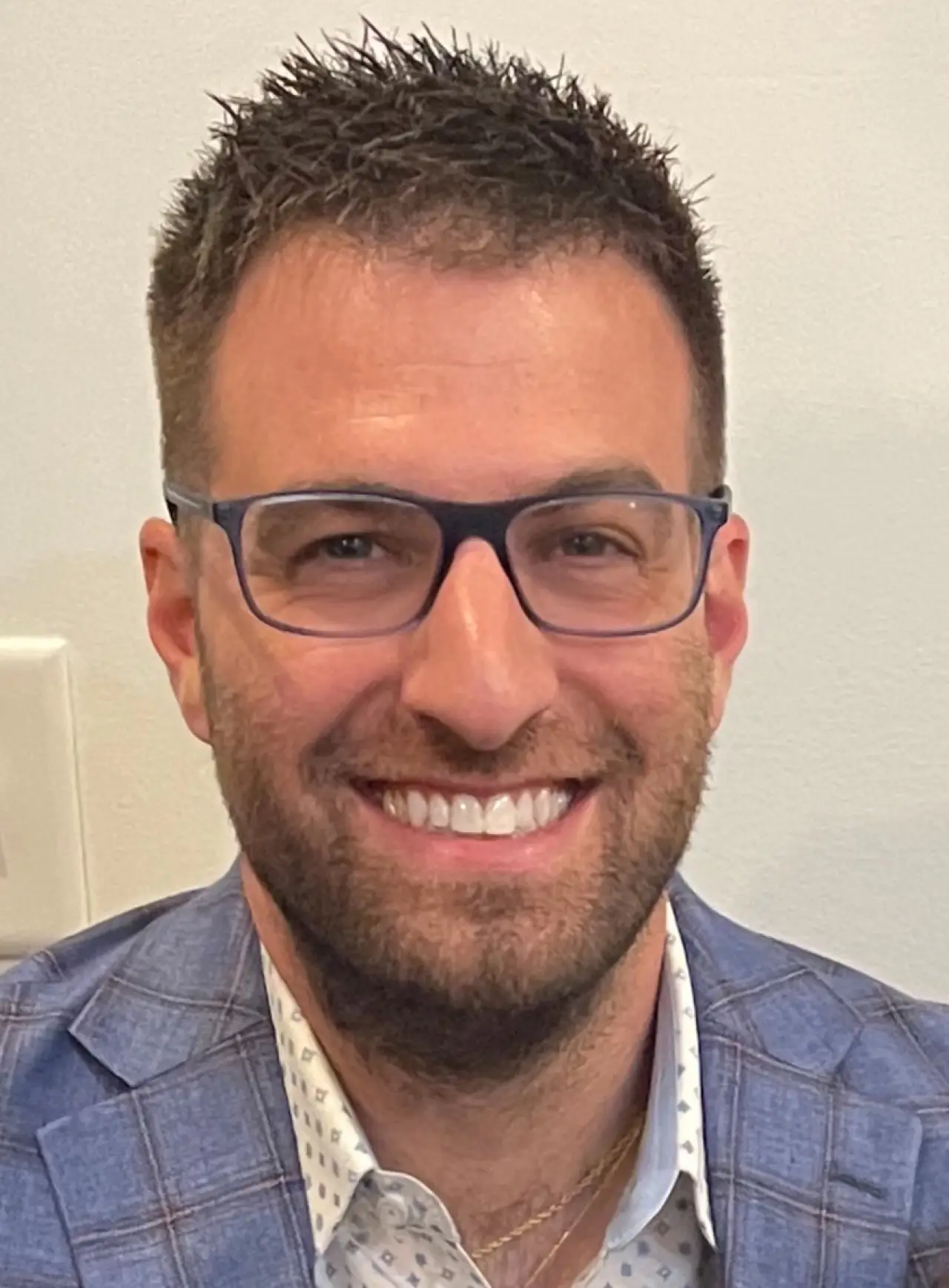 Smiling man with short dark hair, glasses, and a blue blazer against a light background.