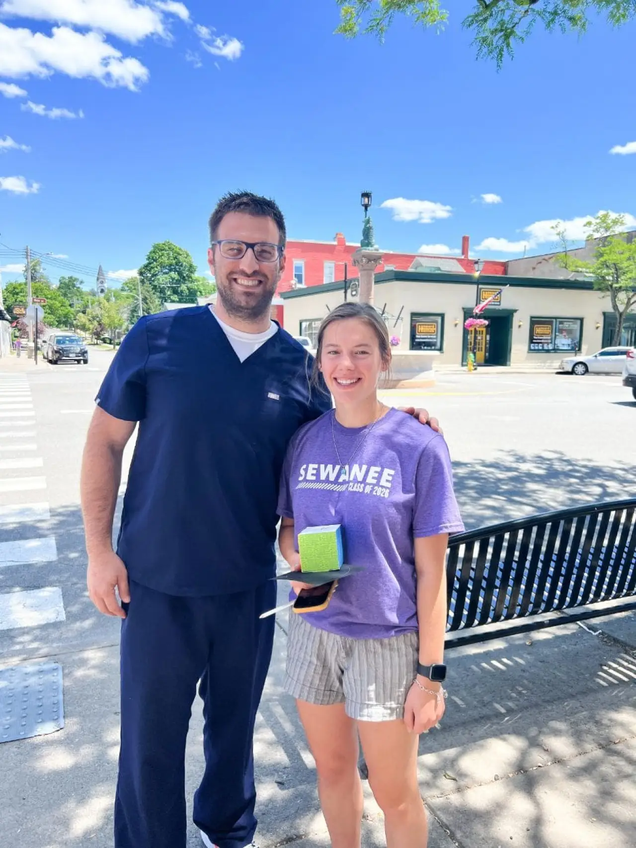 Two people smiling outside; one in navy scrubs, one in a purple Sewanee shirt and striped shorts.