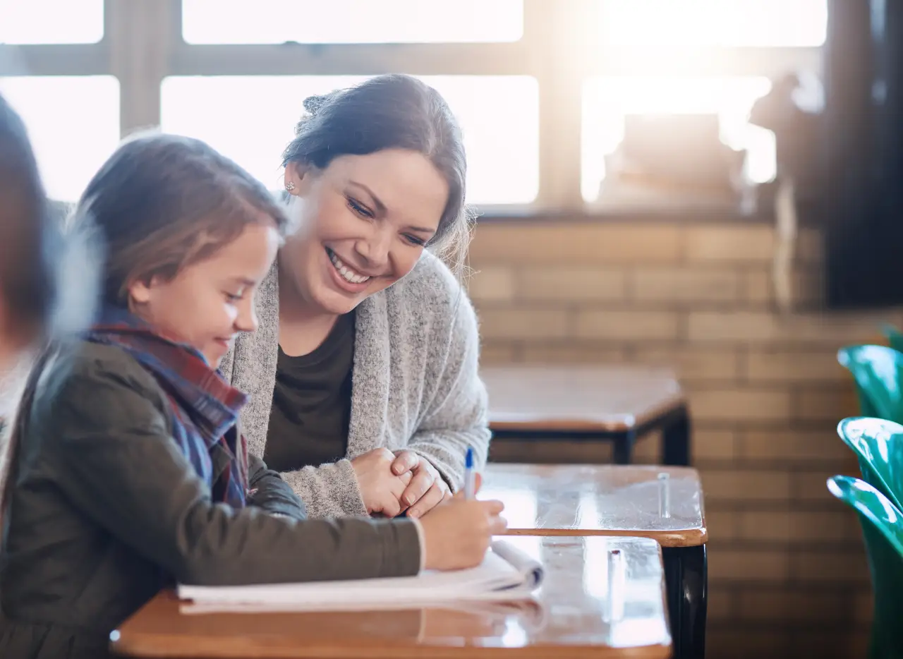 Smiling teacher helps a young student with schoolwork at a classroom desk by a window.