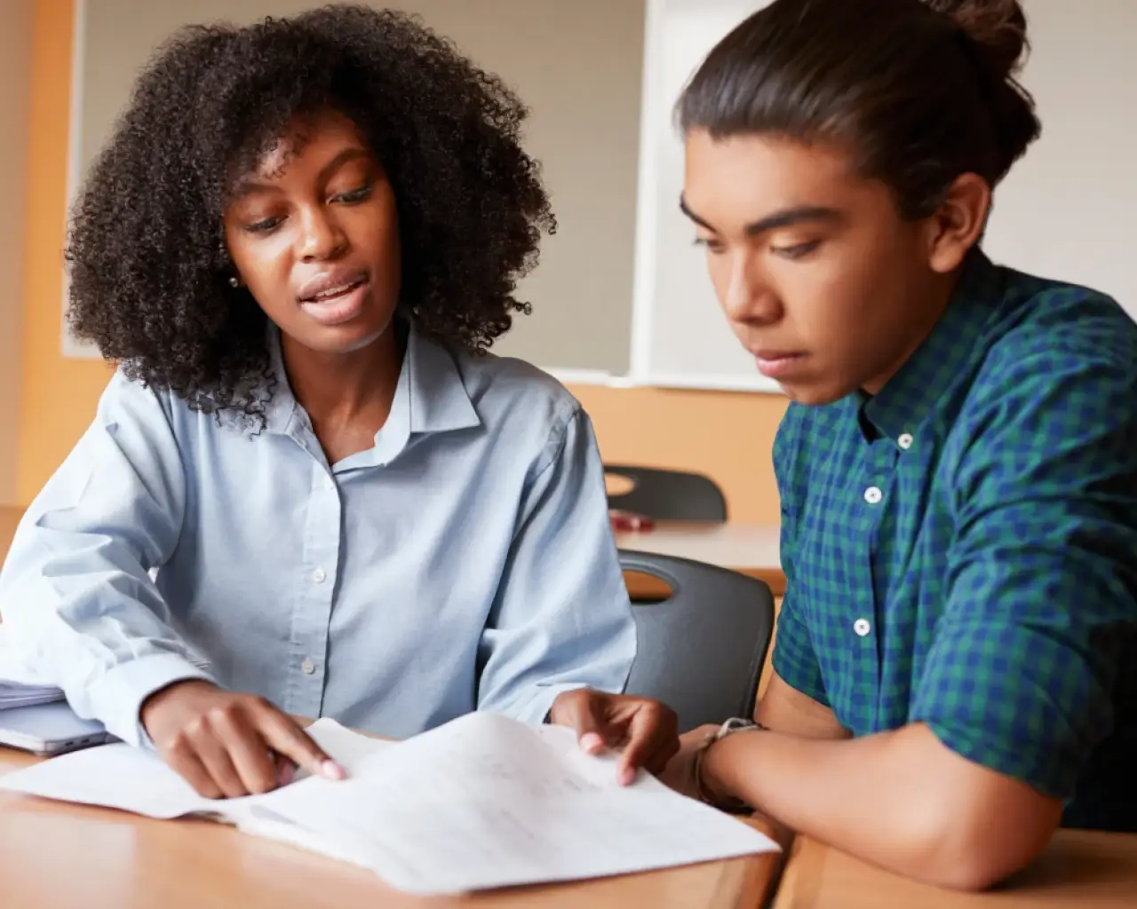 Two students sit at a desk, looking at papers together in a classroom setting.