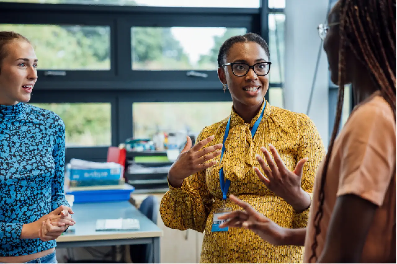 Three women standing and talking in an office, one gesturing with her hands while smiling.