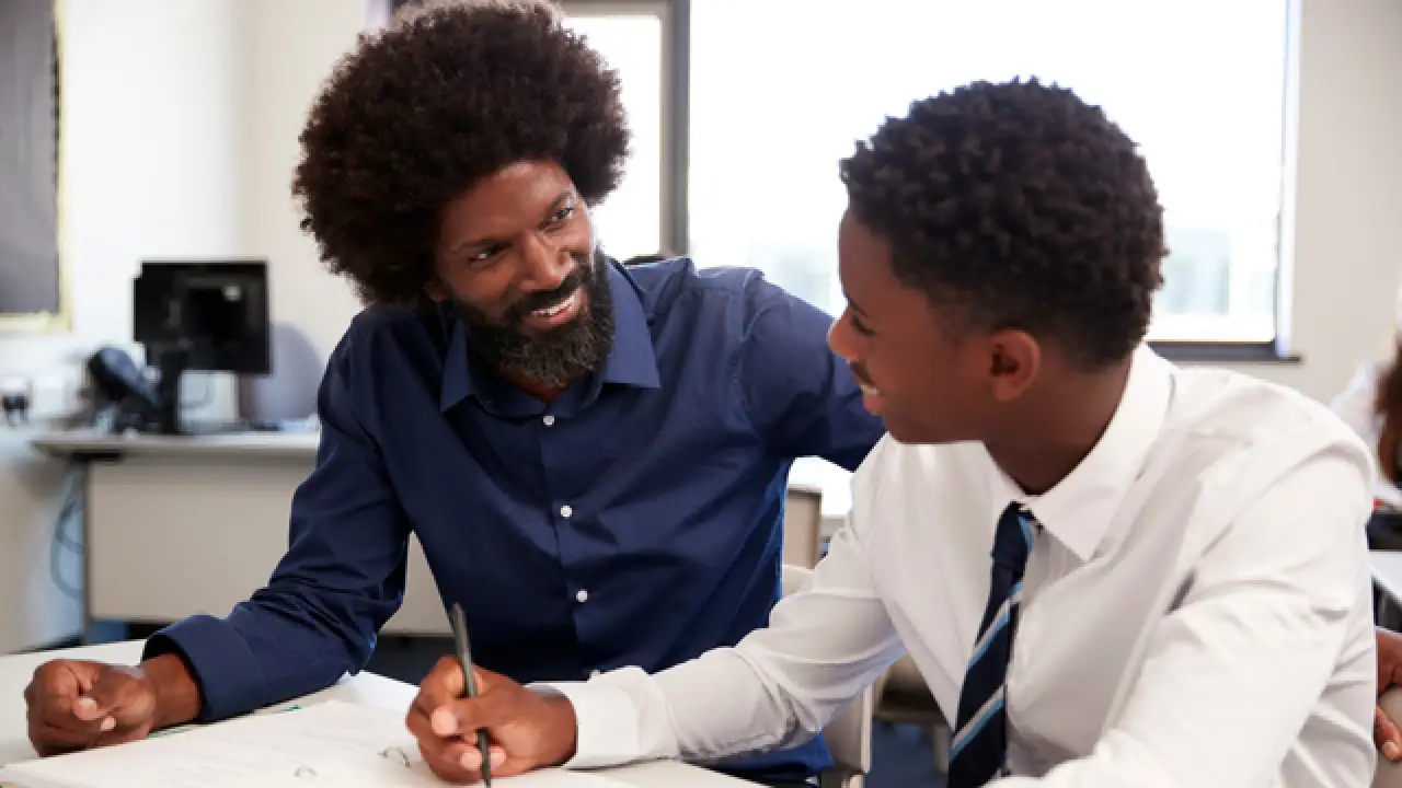 Inside of a classroom, a High- Dosage tutor and high school student sit next to each other at a desk, both smiling at the other.