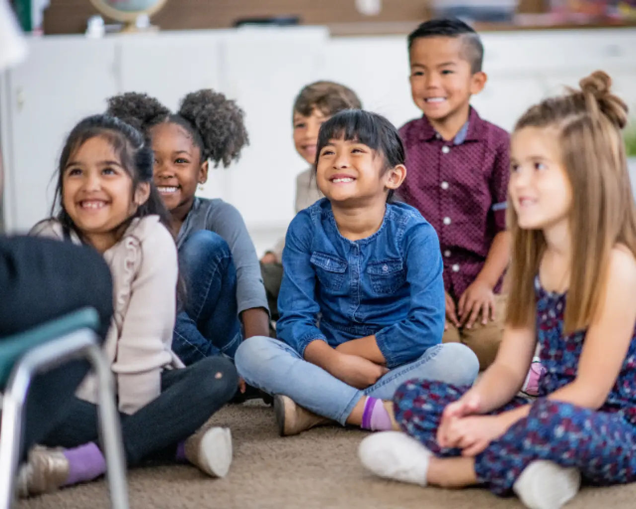 Six first-grade students sit crossed legged on the floor, smiling and facing a teacher sitting on a chair.