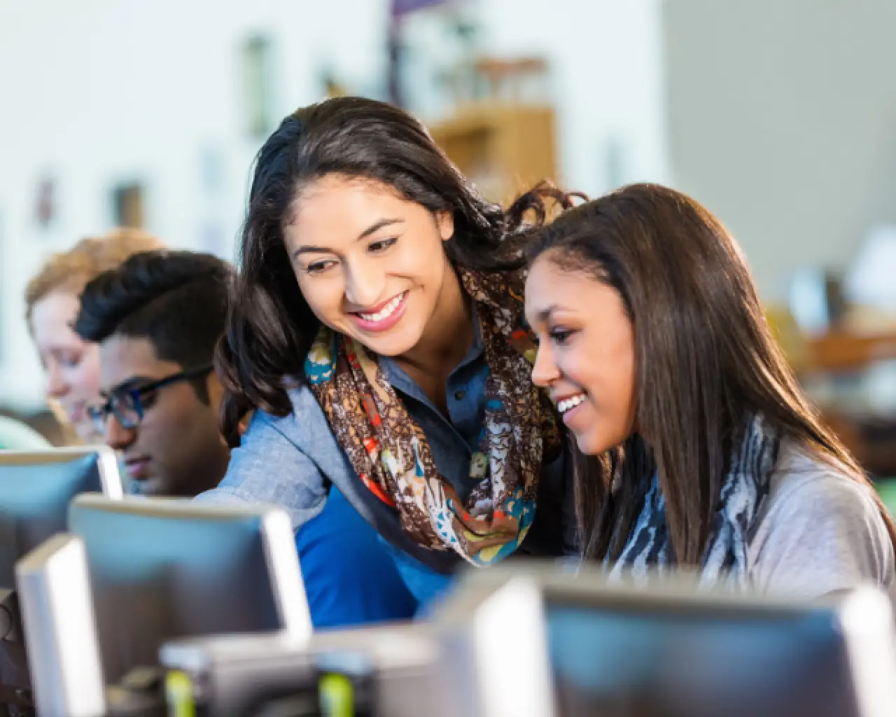 Inside of a computer lab, a smiling High-Dosage tutor stands next to a seated, smiling high- school student. Both are facing toward a monitor, with the tutor pointing at the screen.