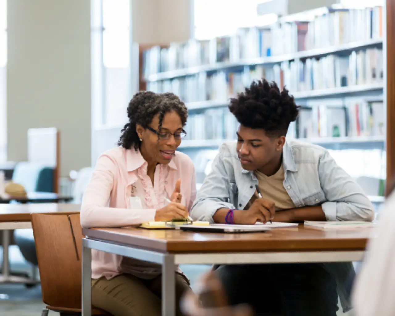 Sitting next to each other at a library table, a High-Intensity tutor points to a notebook with a pen as a high school student looks on.