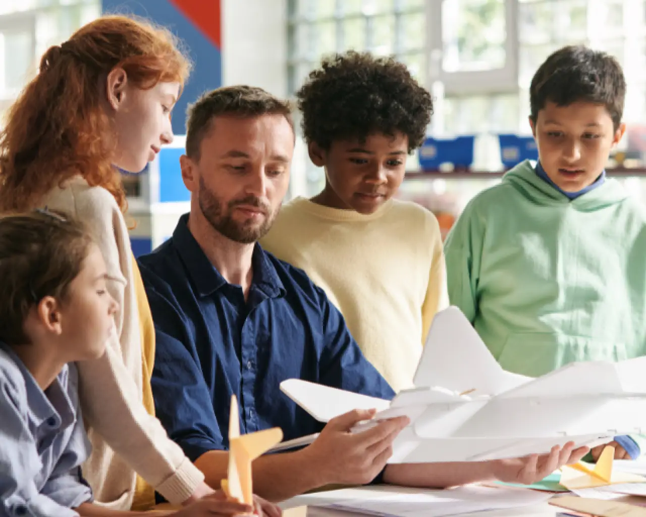 A long-term substitute teacher sits at a desk holding a white- cardboard airplane model, with four students gathered around the teacher looking at it.