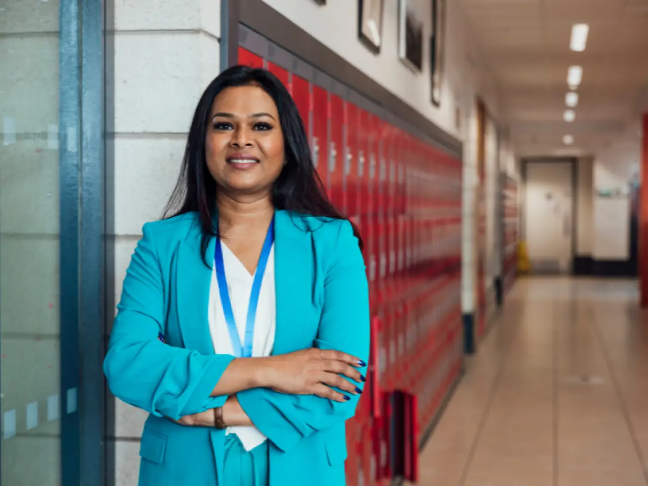 A woman in a blue suit stands confidently with arms crossed in a school hallway by red lockers.