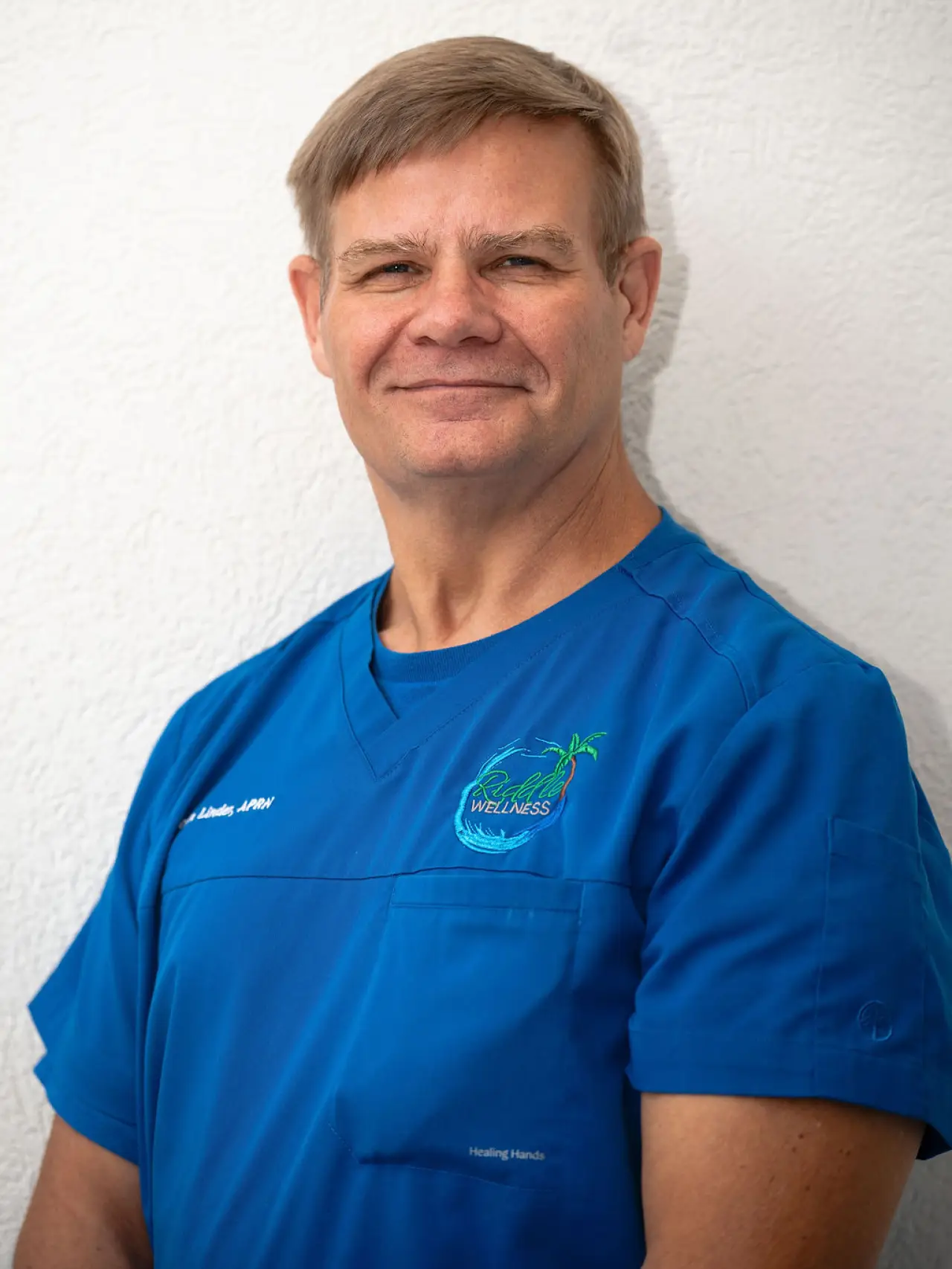 A man in blue medical scrubs stands against a white textured wall, smiling slightly.