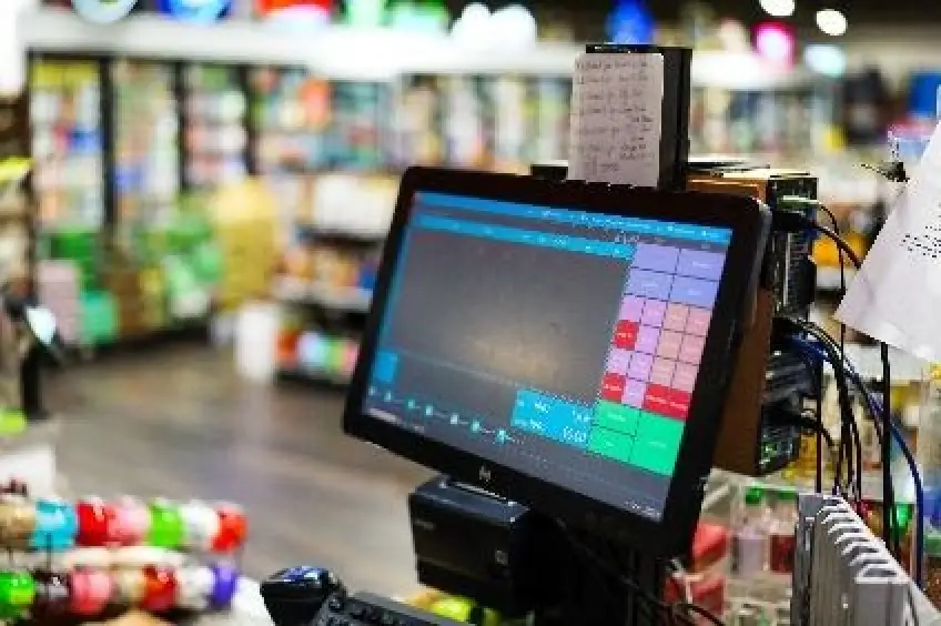 The image depicts a supermarket checkout counter with a computer terminal, cash drawer, and a shopping cart filled with various products, including beverages and tobacco items.
AI-generated content may be incorrect.