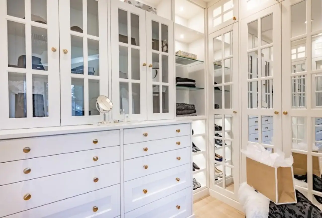 Bright walk-in closet with white cabinets, glass doors, shelves, and a shopping bag on the floor.