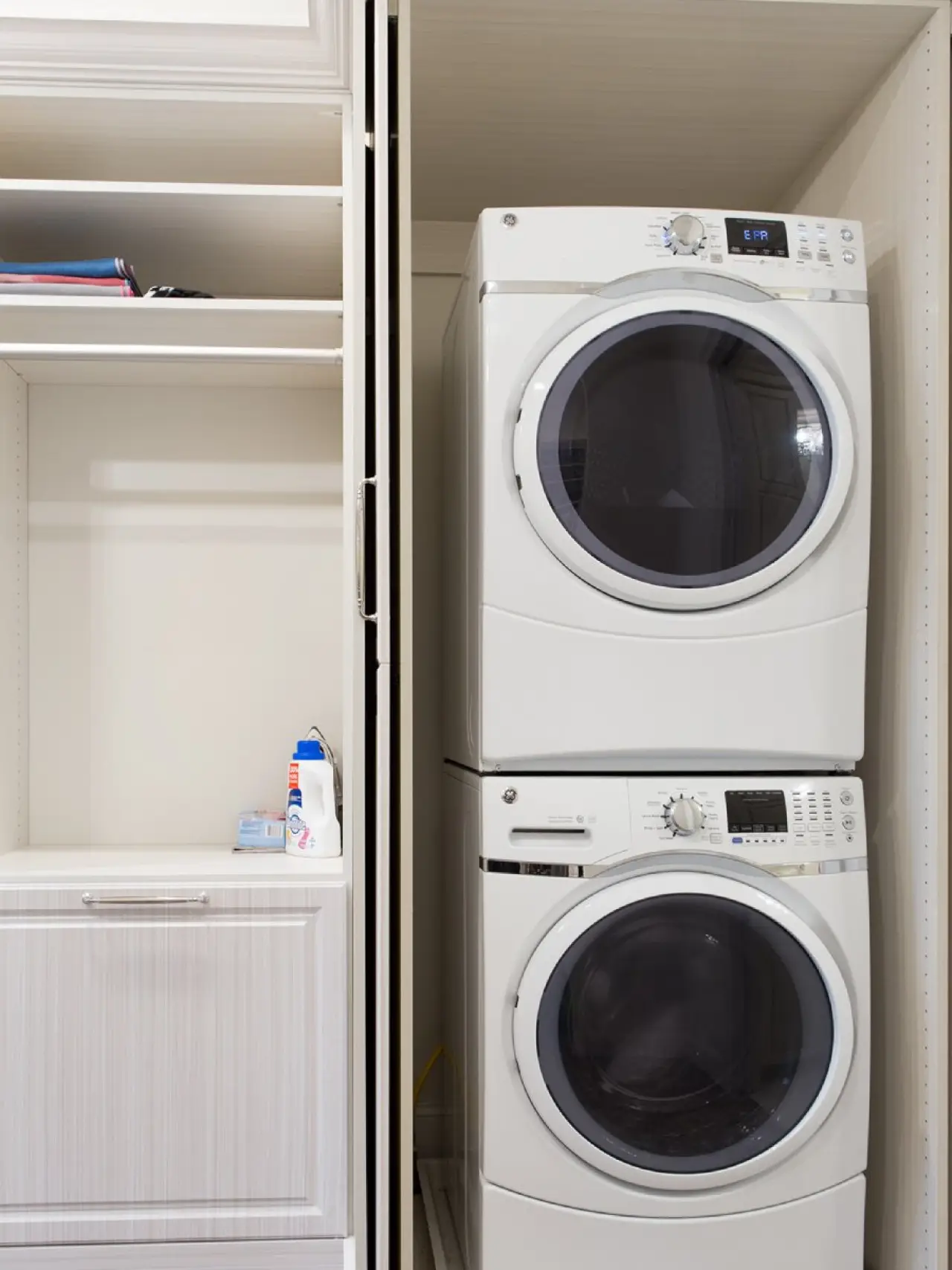Stacked washer and dryer next to shelves and a cabinet in a laundry closet.