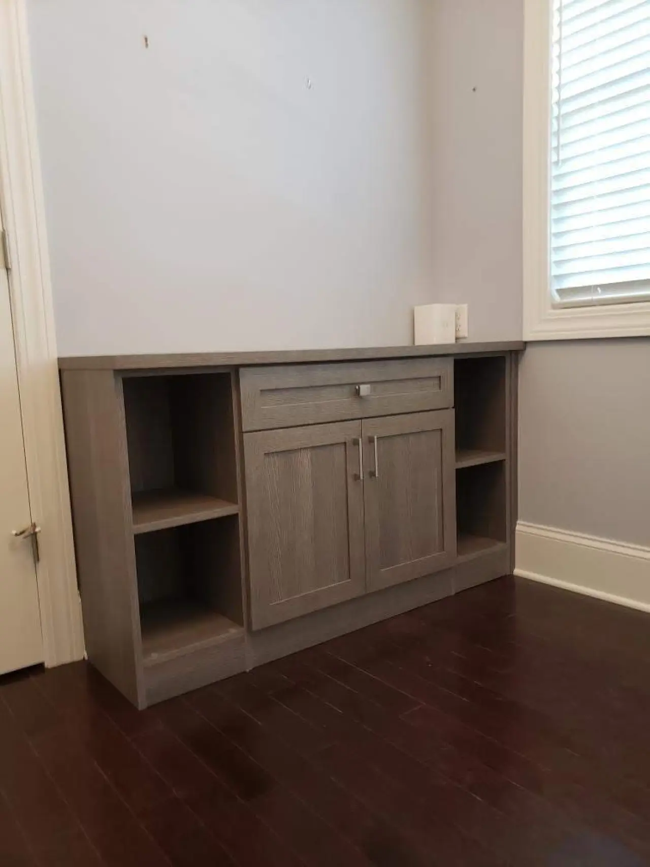 Gray wooden cabinet with open shelves stands against a light wall near a window and closed door.