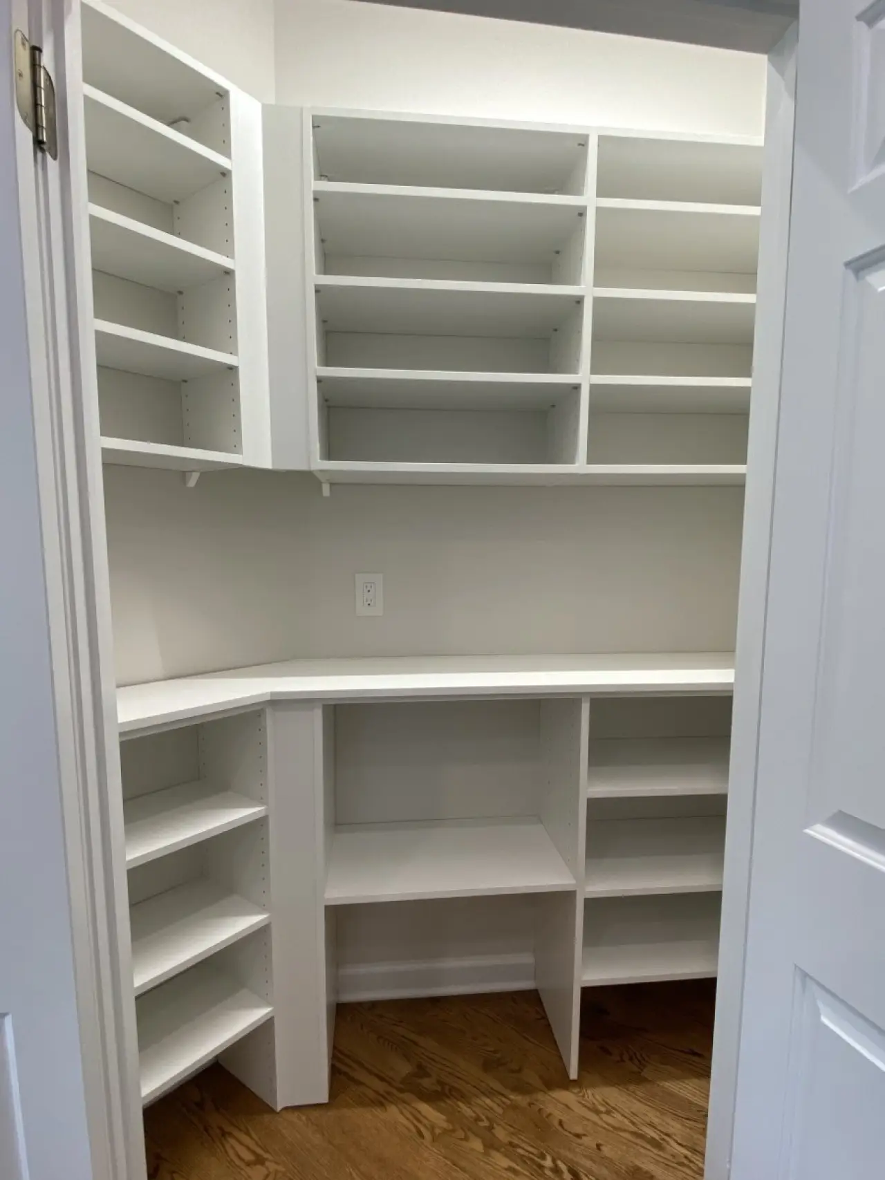 White pantry with empty shelves and hardwood floor, viewed through an open doorway.