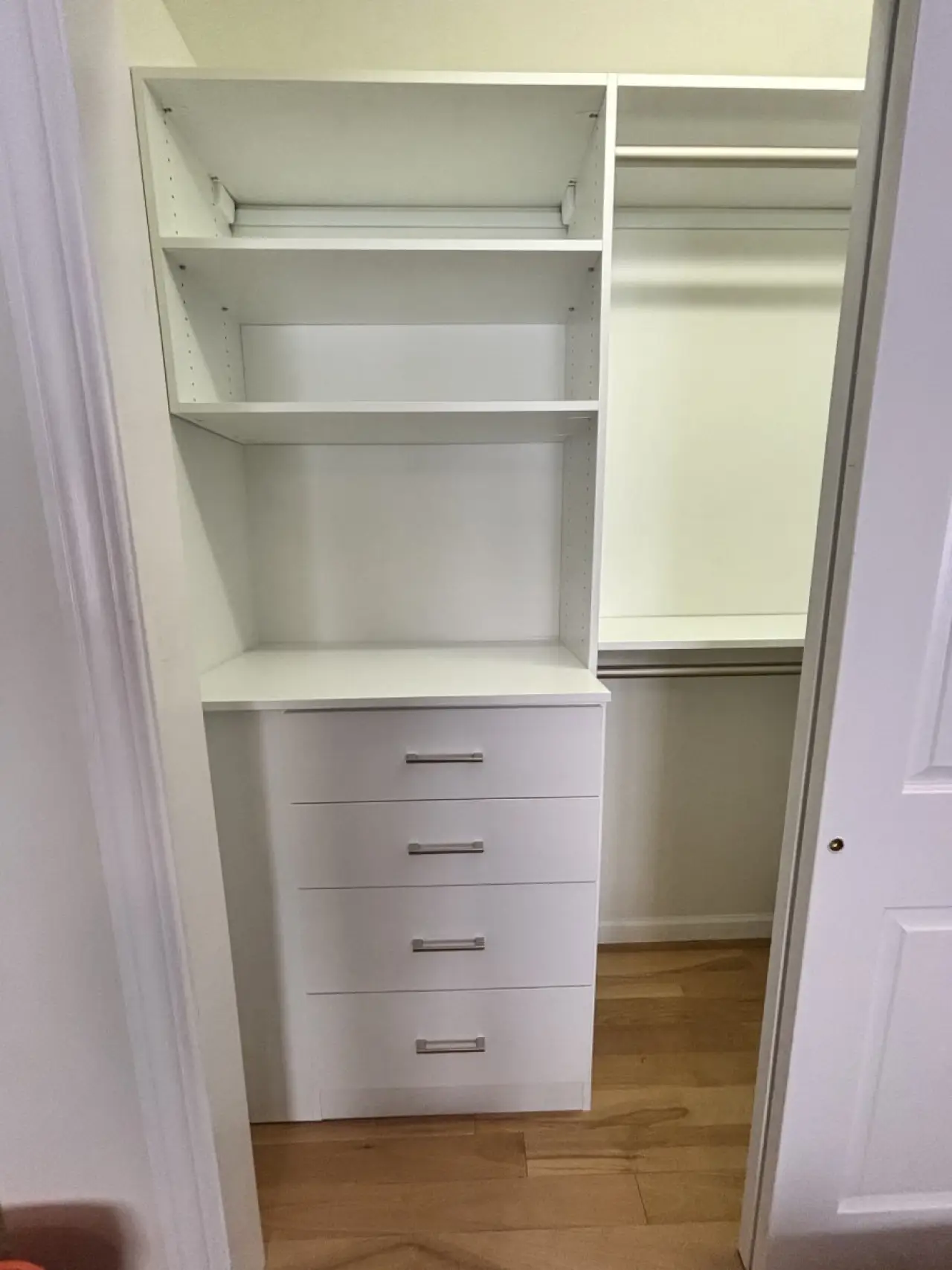 White closet with shelves, drawers, and hanging rod on a wooden floor, viewed from doorway.