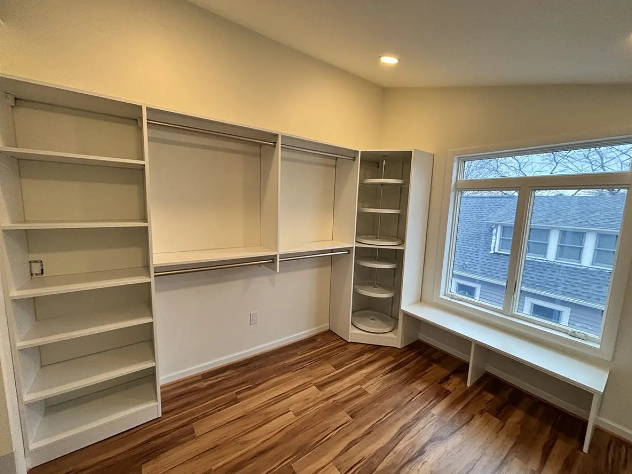 A walk-in closet with white shelves, hanging rods, corner shelves, and a window bench.