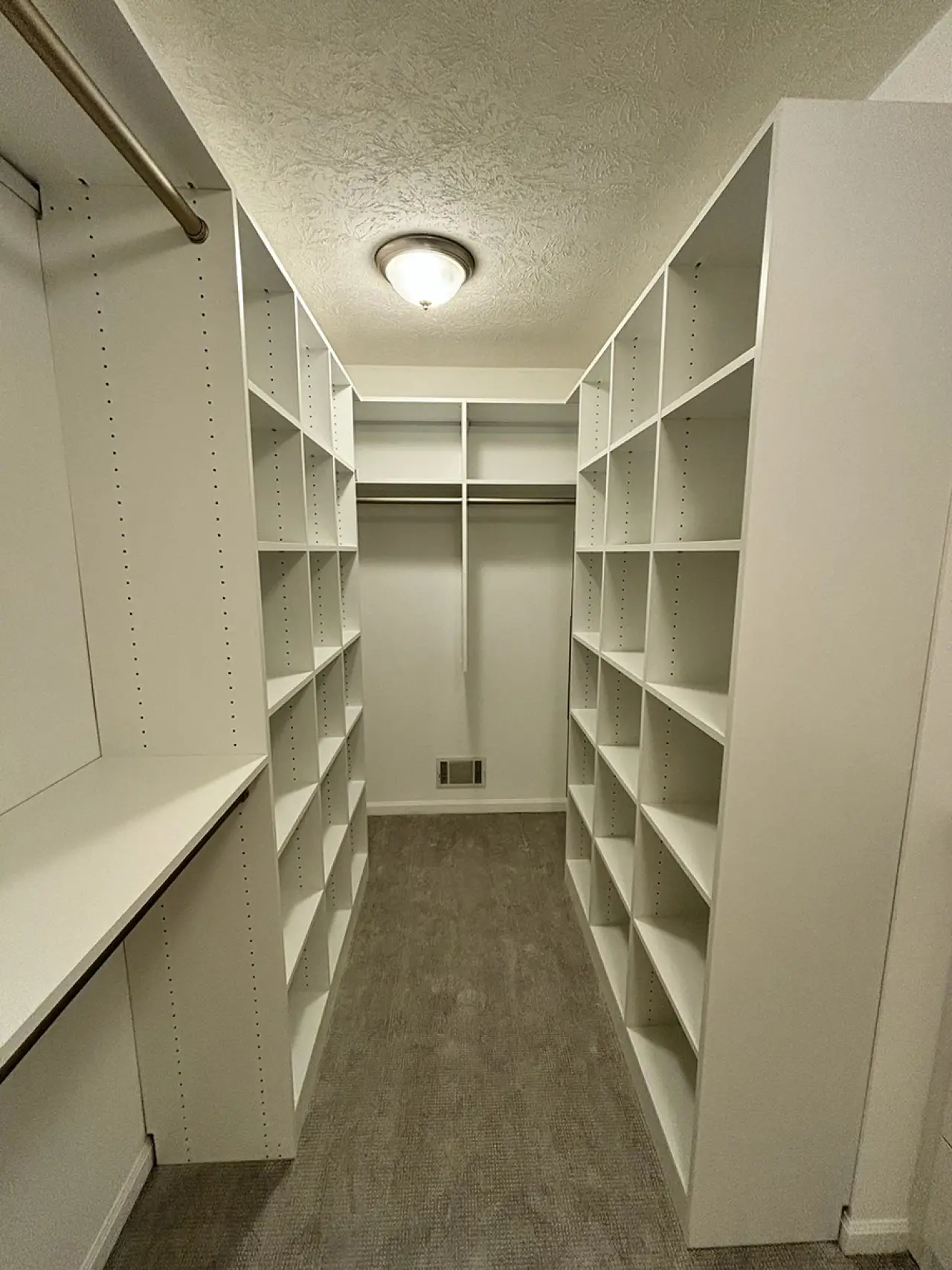A walk-in closet with white shelves, hanging rods, and gray carpet under a ceiling light.