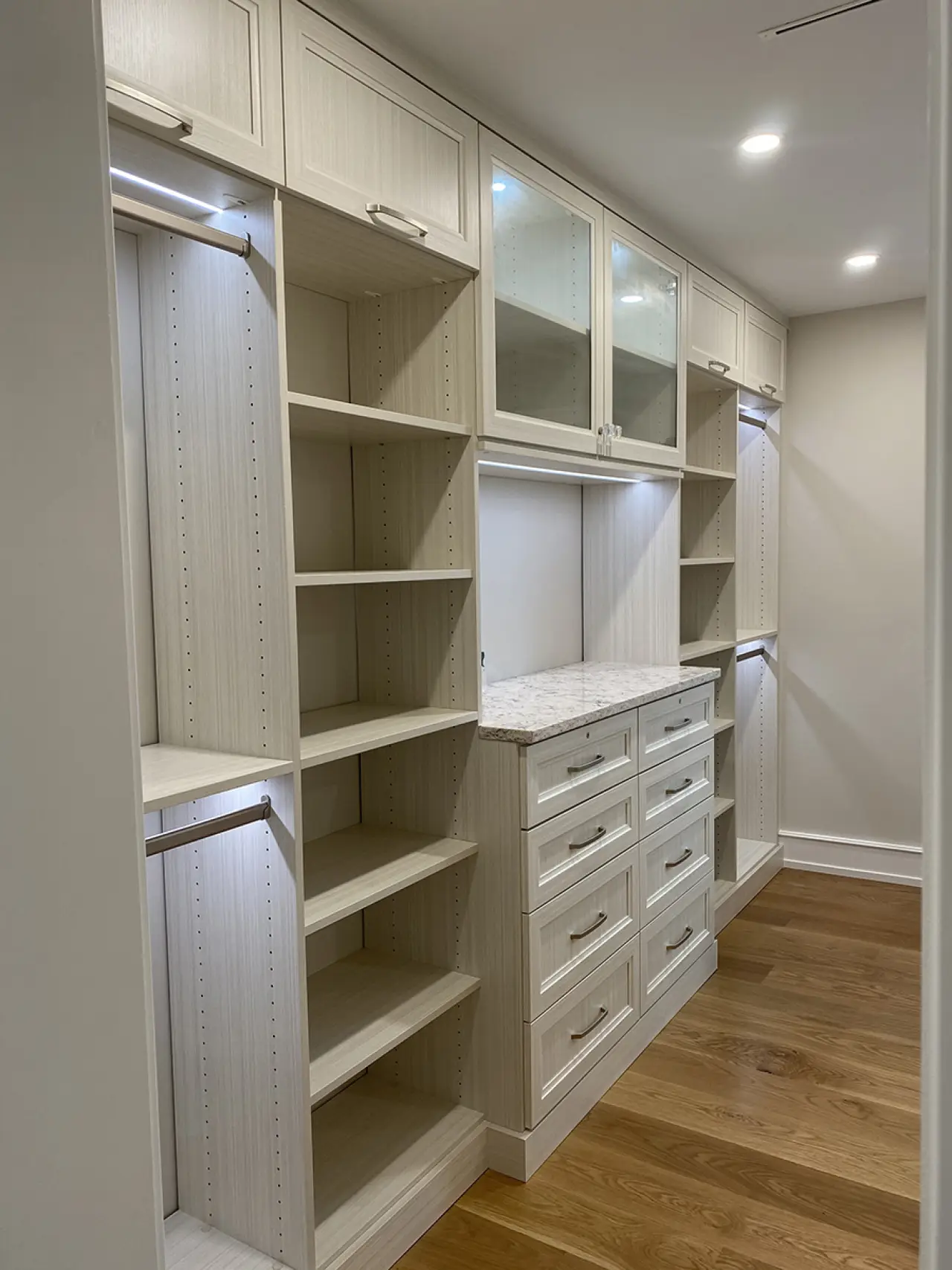Walk-in closet with white shelves, drawers, glass cabinets, and wooden floor, well-lit.