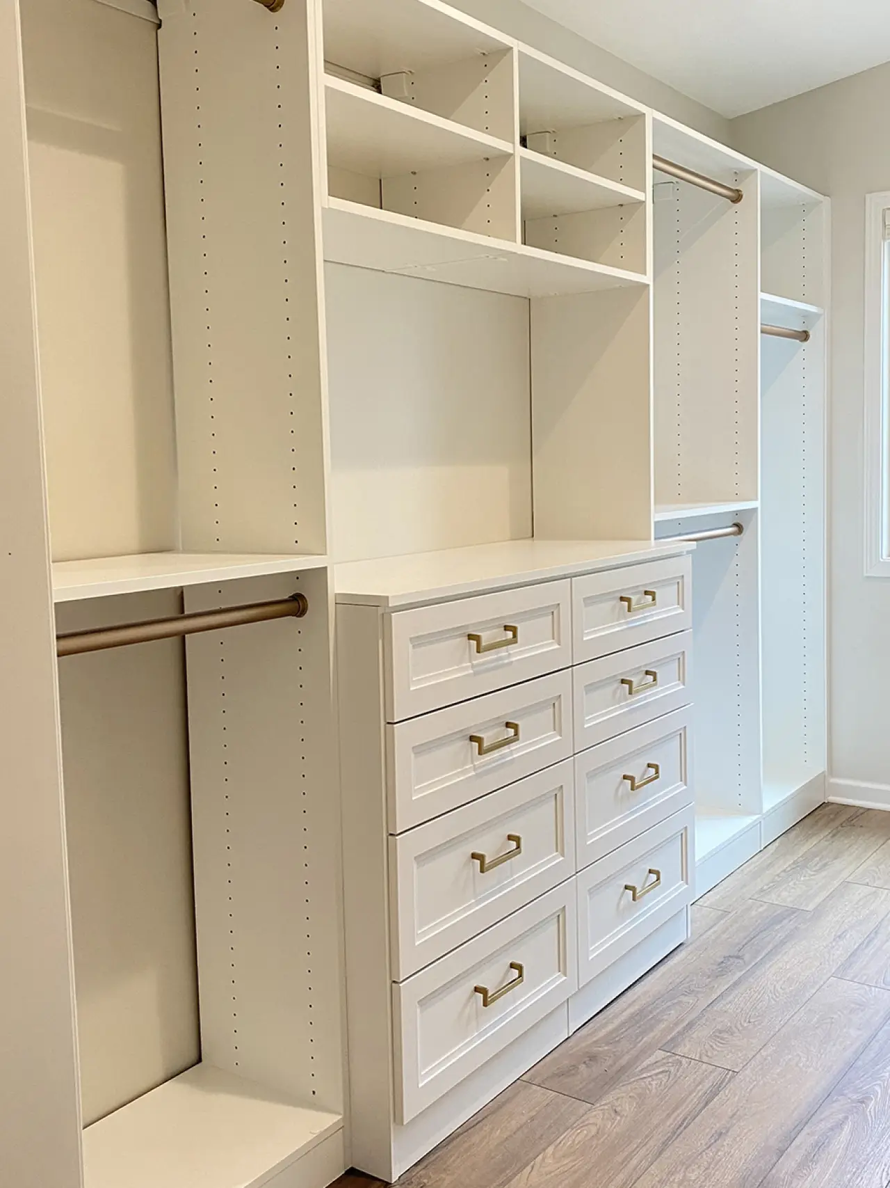 White walk-in closet with gold handles, drawers, shelves, and hanging rods on wood flooring.